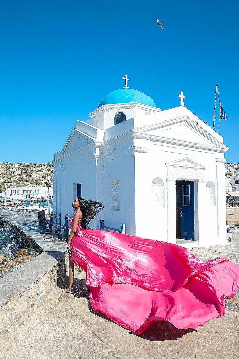 Sunlit fuchsia flying dress at Mykonos blue dome chapel at a Mykonos windmill, photography in Mykonos, Greece