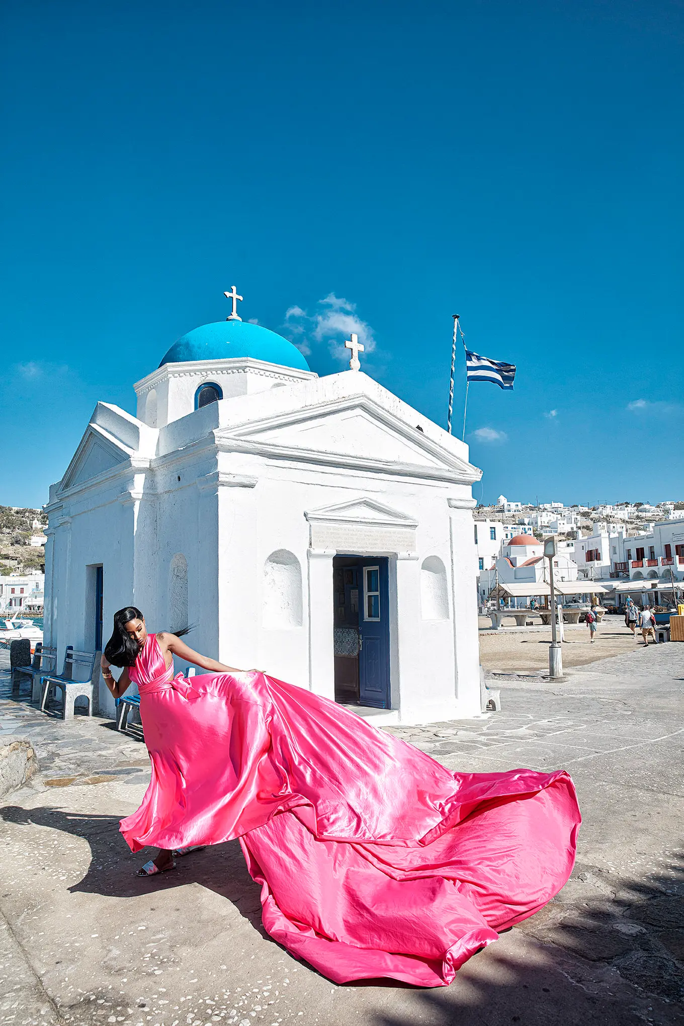 Sunlit fuchsia flying dress at Mykonos blue dome chapel on a Mykonos rooftop, photography in Mykonos, Greece