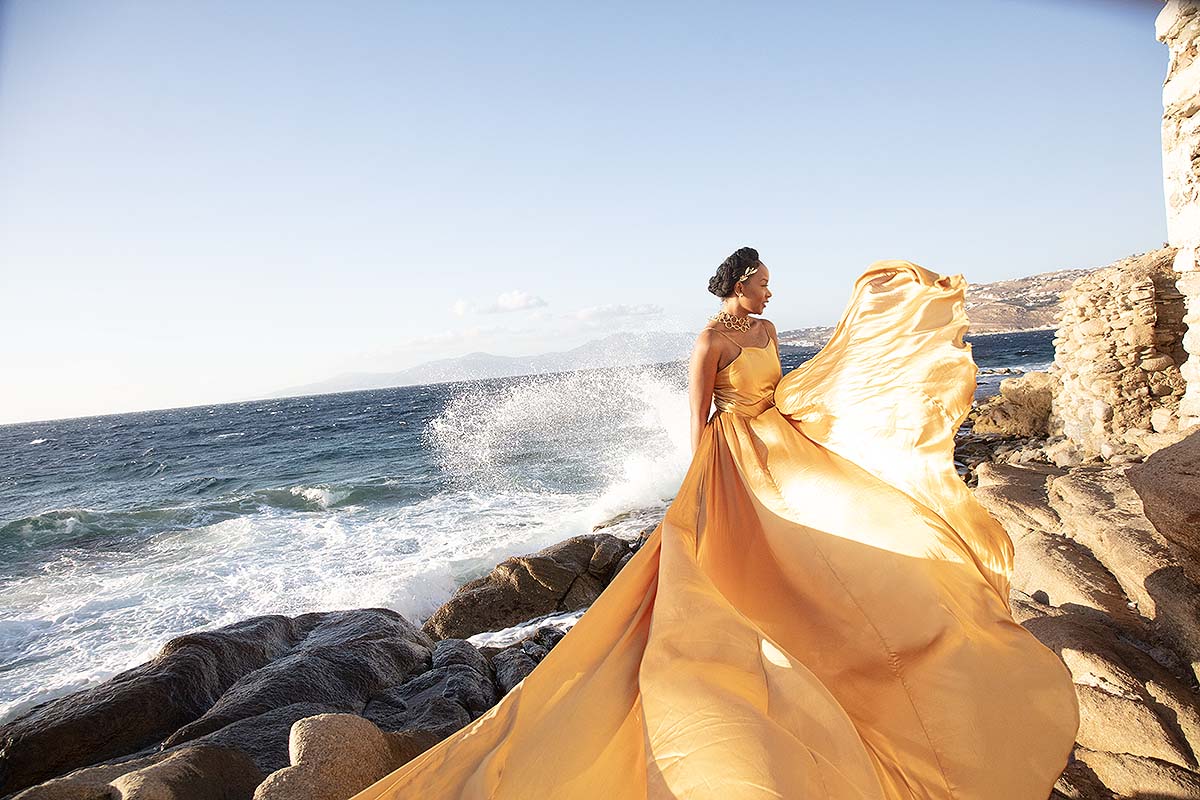 Sunlit red satin flying dress at Mykonos windmill in a Mykonos alley, photography in Mykonos, Greece
