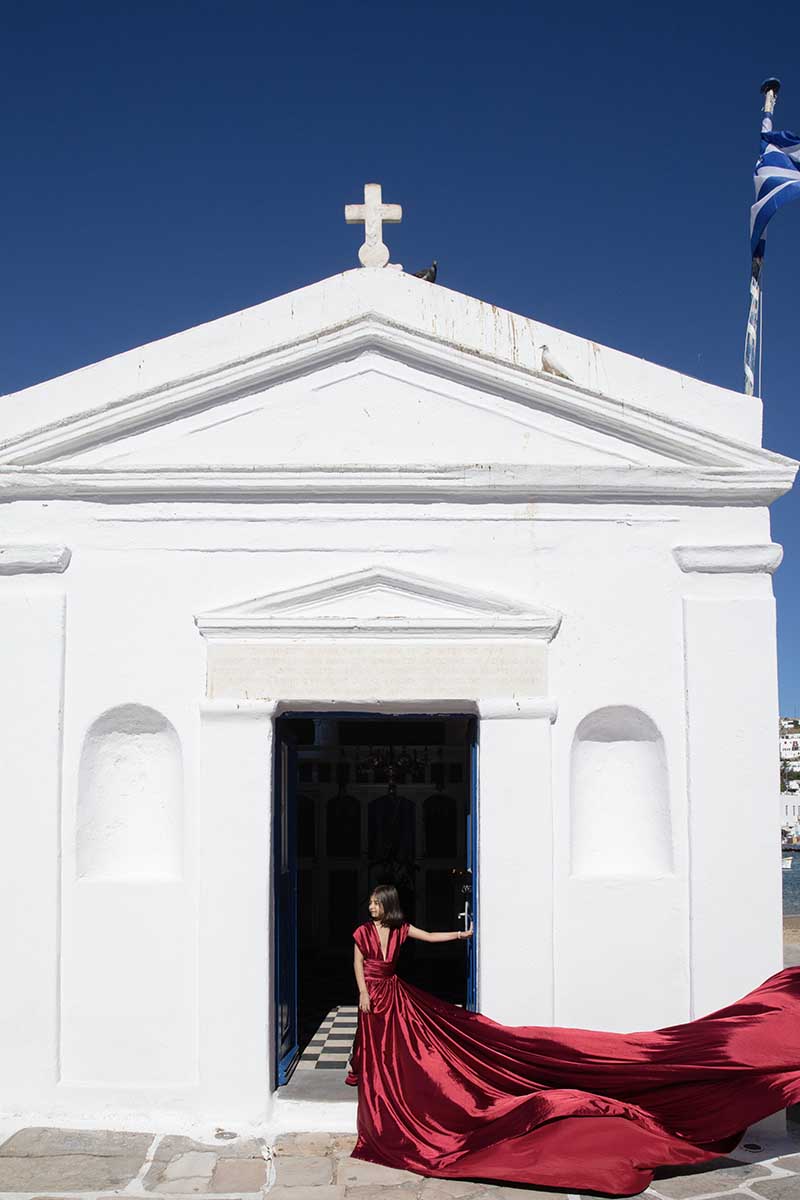 Sunlit red satin flying dress at Mykonos windmill on a Mykonos rooftop, photography in Mykonos, Greece