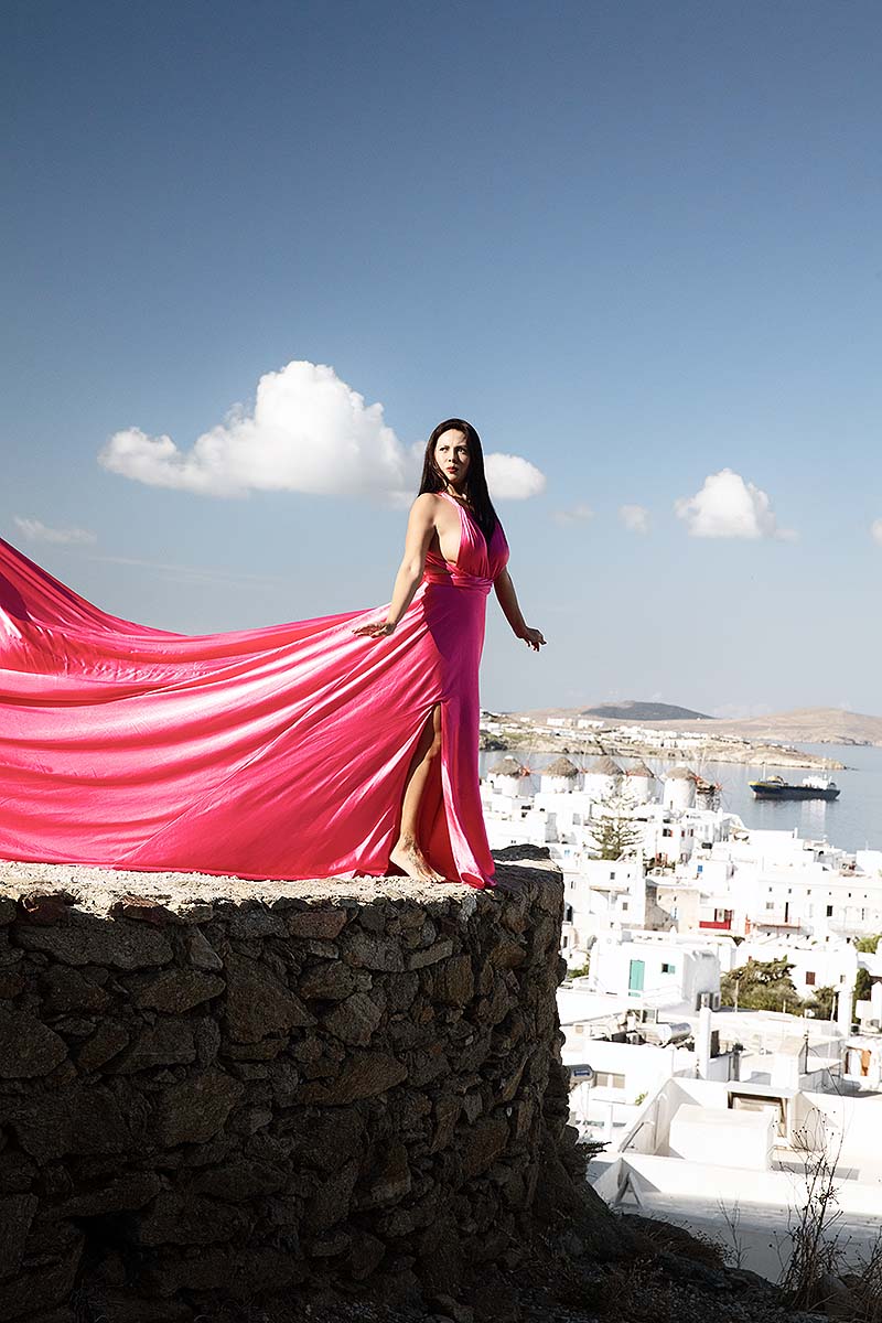 Sunlit red satin flying dress at Mykonos windmill by Cycladic walls, photography in Mykonos, Greece