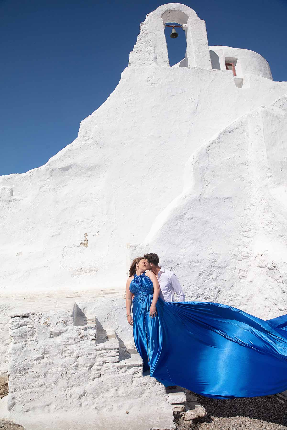 Sunlit red satin flying dress at Mykonos windmill near a whitewashed chapel, photography in Mykonos, Greece