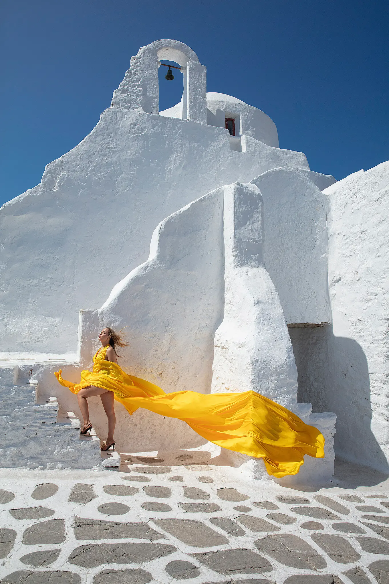 Sunlit teal flying dress near a whitewashed chapel, photography in Mykonos, Greece