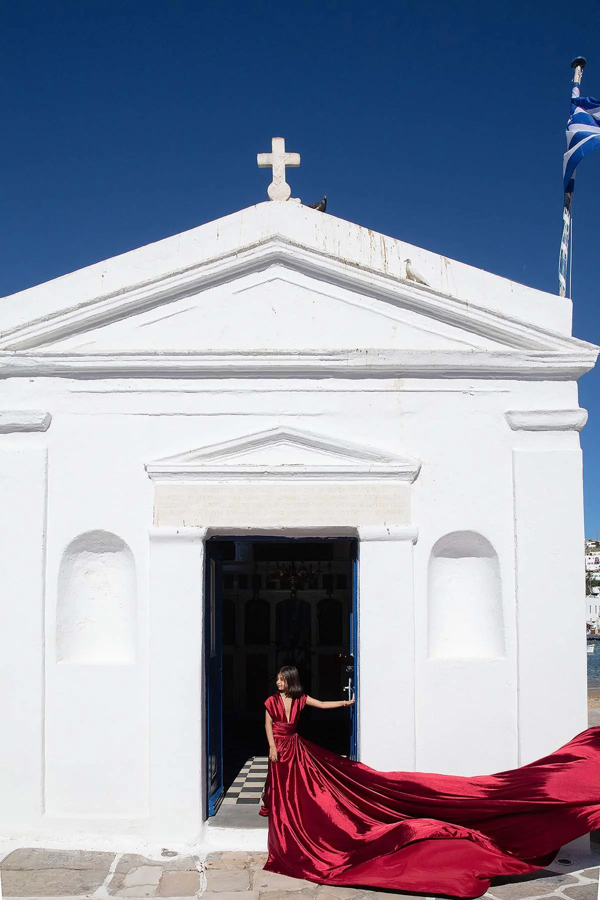 Sunlit red flying dress at white Mykonos chapel at a Mykonos windmill, photography in Mykonos, Greece