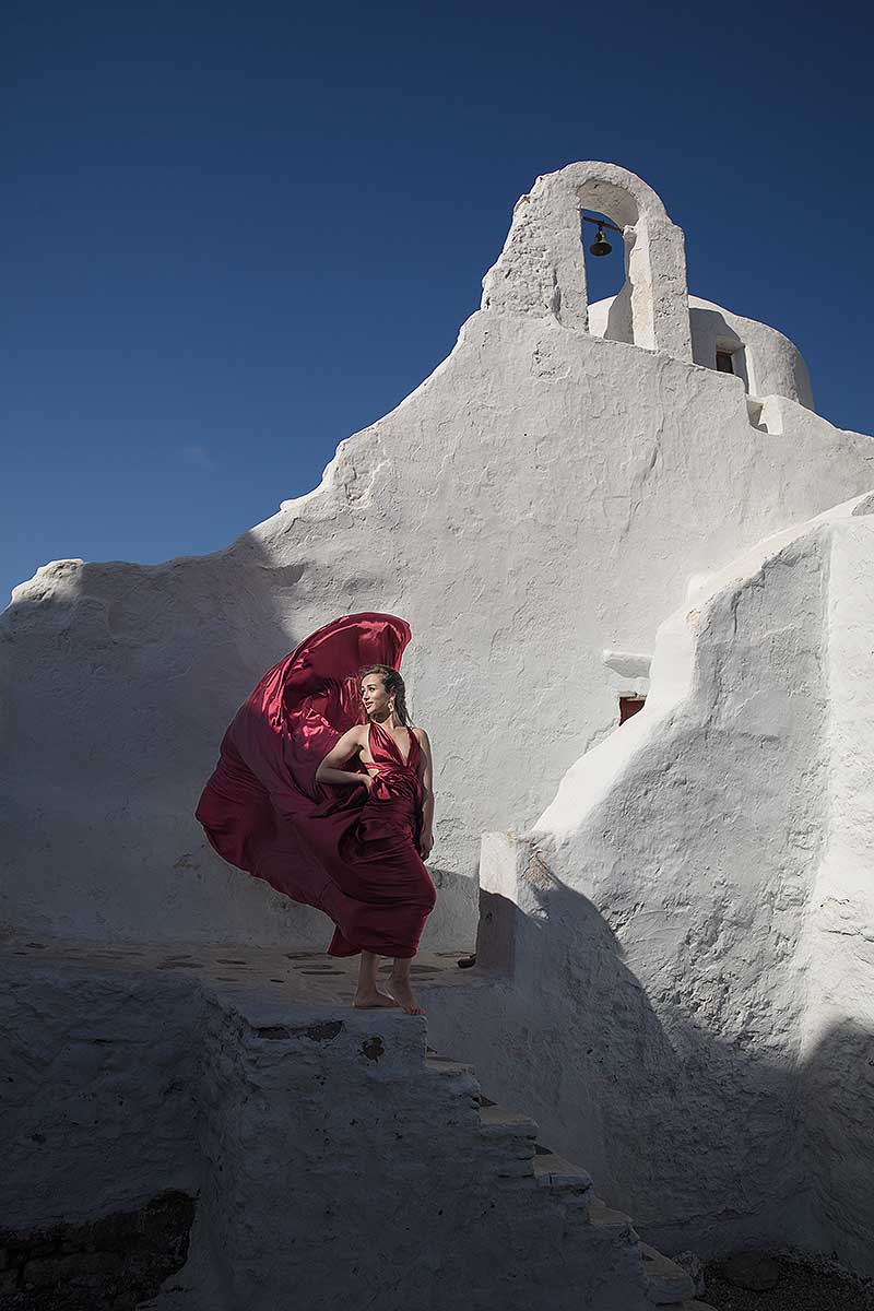 Sunlit red flying dress at white Mykonos chapel by the Aegean sea, photography in Mykonos, Greece