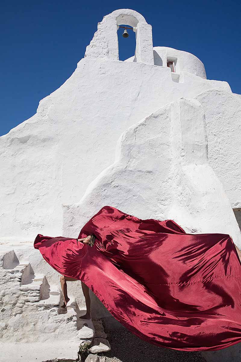 Sunlit red flying dress at white Mykonos chapel near a whitewashed chapel, photography in Mykonos, Greece